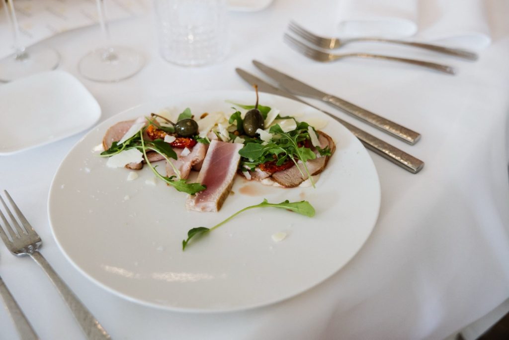 Een wit bord met plakjes gebakken tonijn, rucola, kappertjes en zongedroogde tomaten, gegarneerd met een lichte dressing. Zilverwerk en een gevouwen witte servet liggen op een wit tafelkleed op de achtergrond.